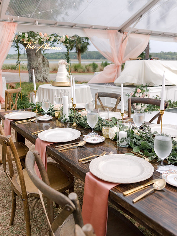A beautiful table set up for dining under a tent. Peach-colored linens and gold flatware with flowers as a runner.